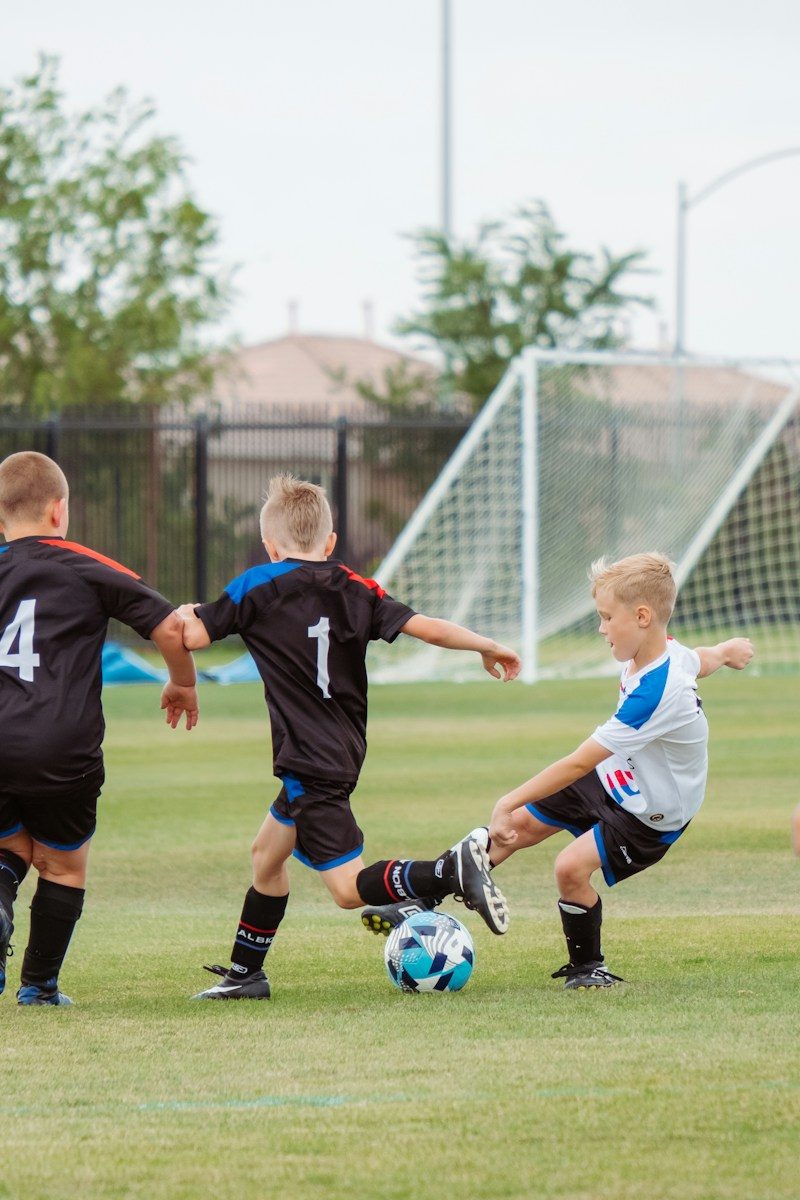 Three children playing soccer on a grass field with a goal in background